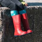 Classic Wellies Red: Red wellington boots on a concrete ledge, worn with teal socks and black trousers. A hand rests nearby.